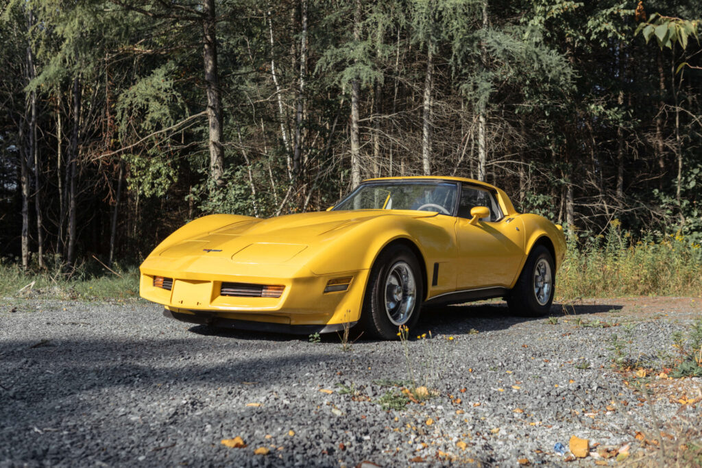 Full view of a 1980 yellow Corvette with a forest in the background