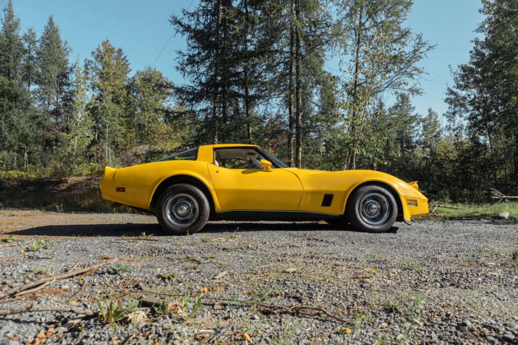 Yellow 1980 Corvette in profile with open head lights. The road is made of gravel and green trees are seen in the background.