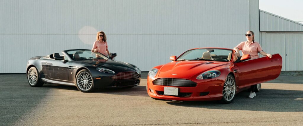 Two Aston Martins are parked facing each other in diagonal. Two girls are standing by their side