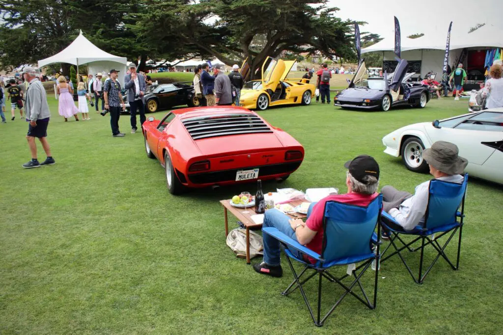 Two individuals are sitting on folding shares looking at a red Lamborghini Miura at the Concorso Italiano