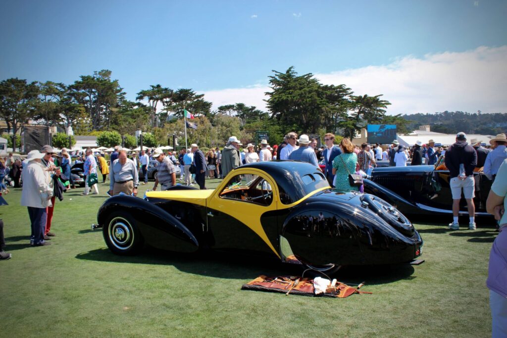 Same black and yellow Bugatti seen earlier but stationary in the crowd at the Pebble Beach Concours d'Elegance 2023. A layer out toolkit is by its side.