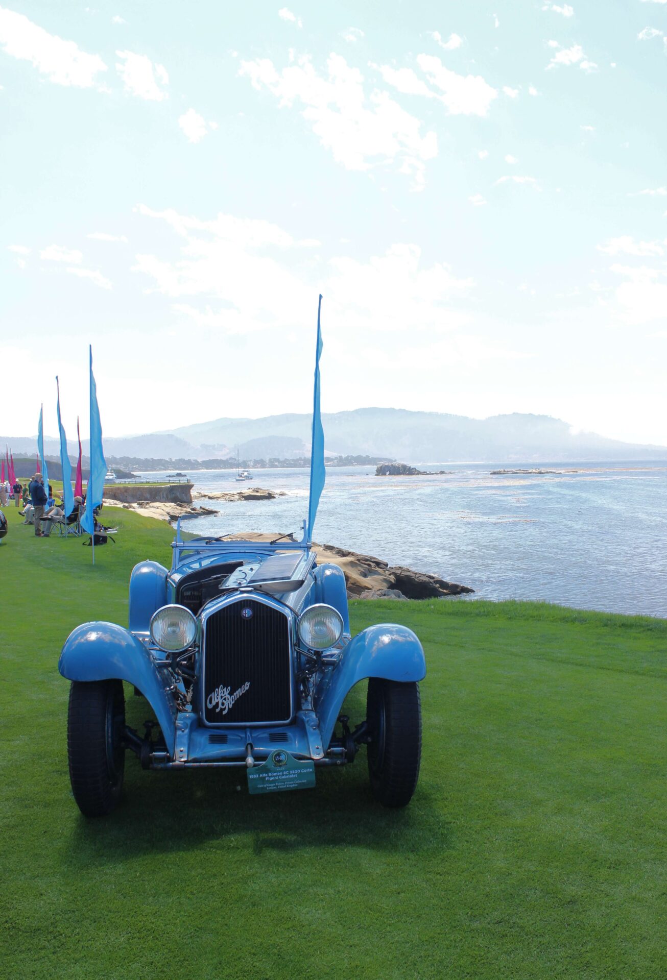 Blue Alfa Romeo from view at the Pebble Beach Concours d'Elegance 2023. Car is on green grass in front of the water, mountains seen in the back. A row of blue flags lines the water.