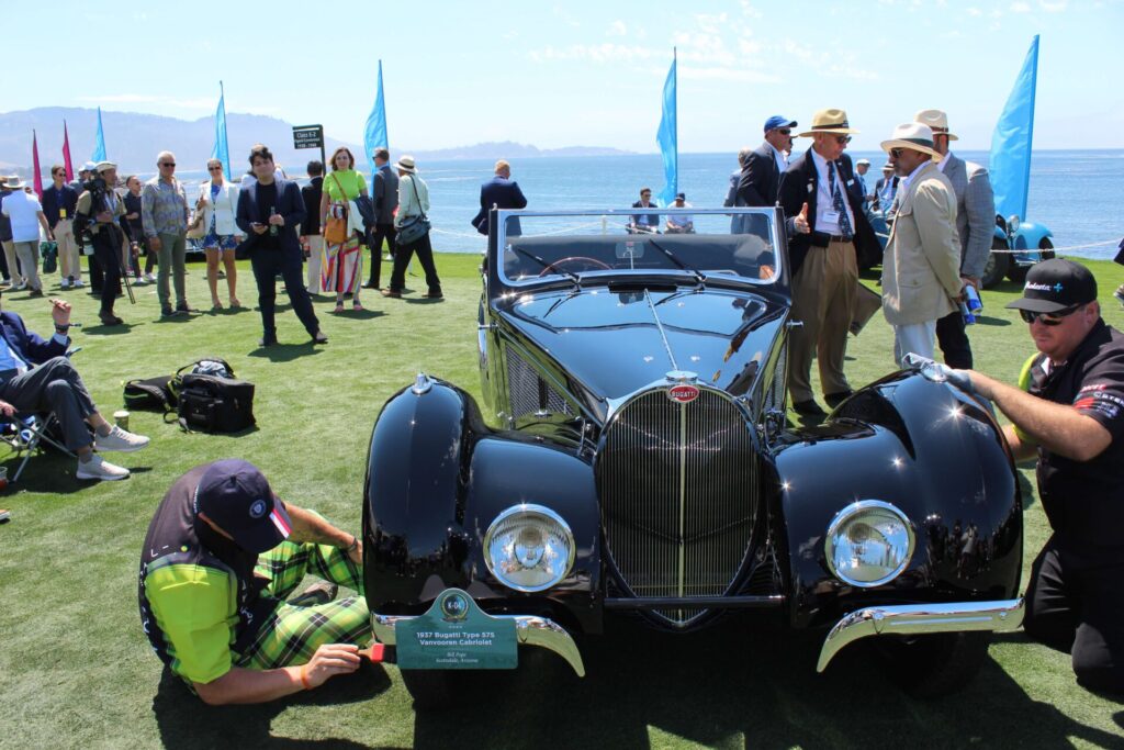 Two individuals in marching black and green outfits are working on polishing a black Bugatti car. One is working on the tire with a brush while the other is working on the wing with a cloth