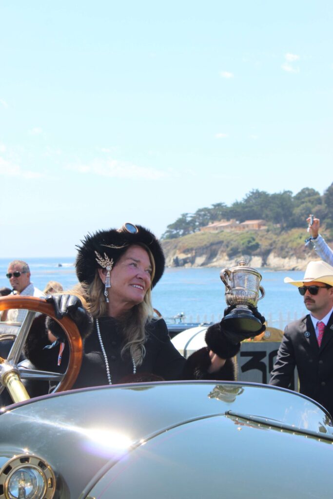 Lady in era costume holding a Cristal prize. She is seated behind the wheel of a bronze era car