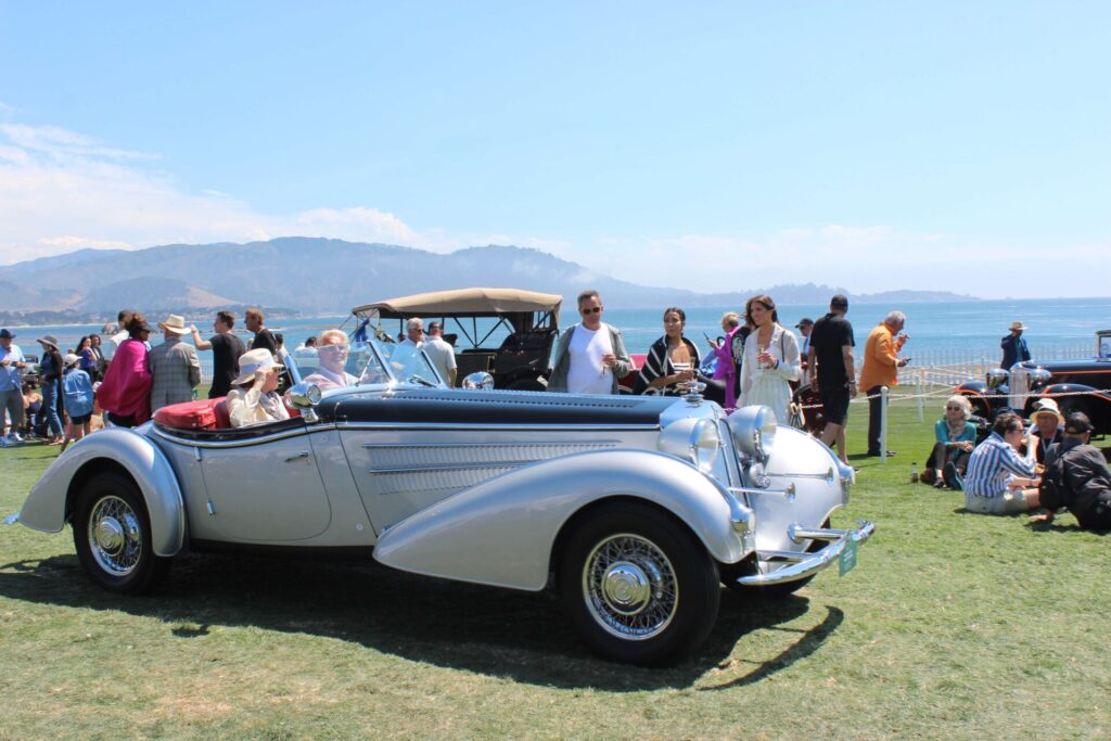 Two individuals riding in a  Horch through the crowd at Pebble Beach Concours d'Elegance 2023