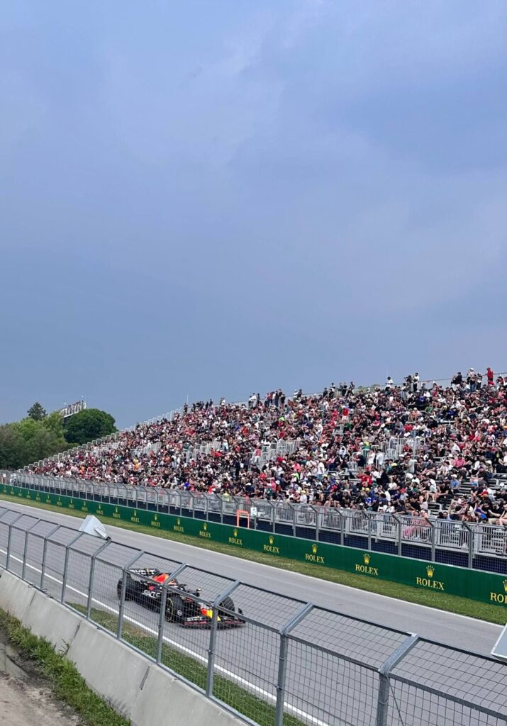 An F1 racing car on the Gilles-Villeneuve circuit as seen from a spectator. Bleachers in  the background