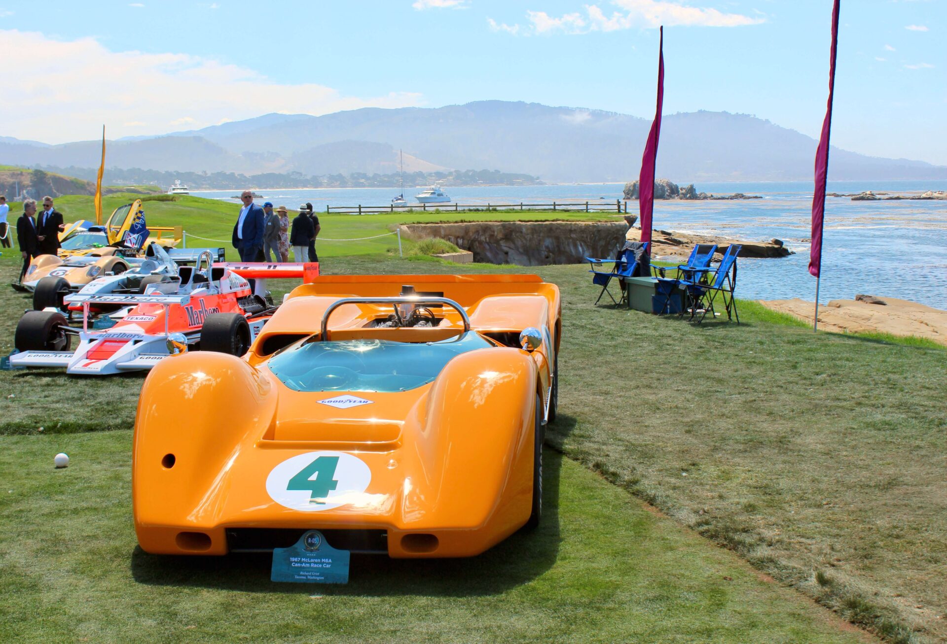 An orange racing car is in the forefront of the frame. It is the first of a row of other racing cars of all different shapes. The setting is on the grass next to a rocky beach.