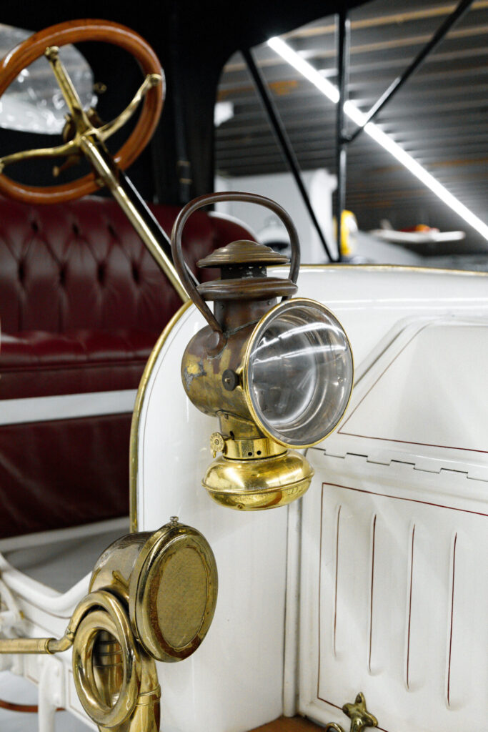 Close up of the bronze lantern on a 1900s white car. Part of the horn, also in bronze can be seen as well as the steering wheel, also partly in bronze.