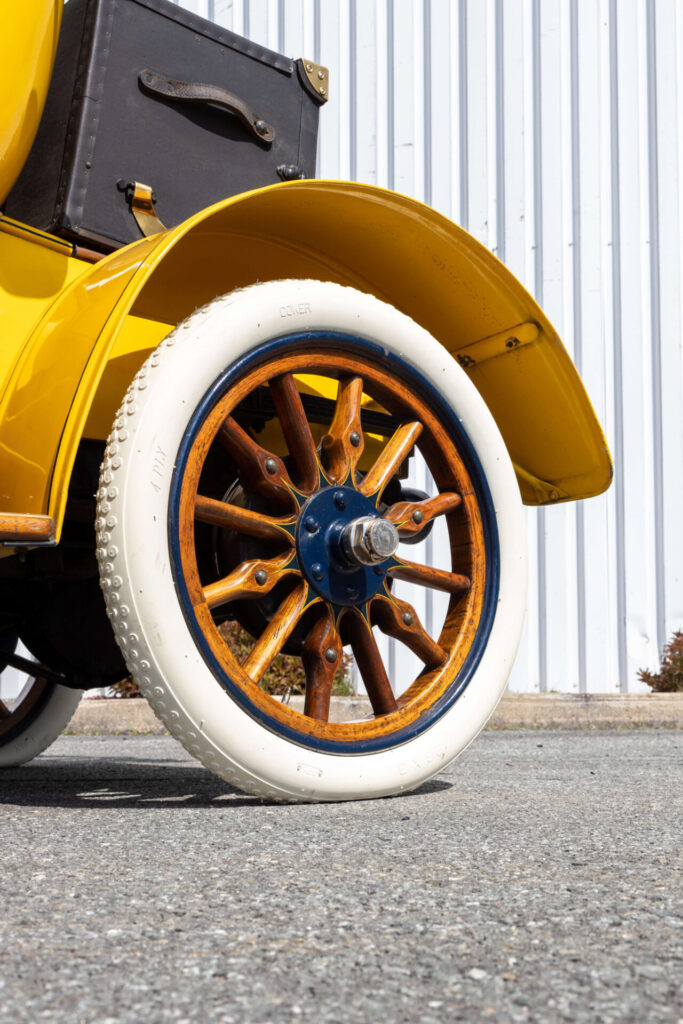 Gros plan sur une roue à rayons en bois sur une voiture jaune au look ancien. Le pneu est entièrement blanc.