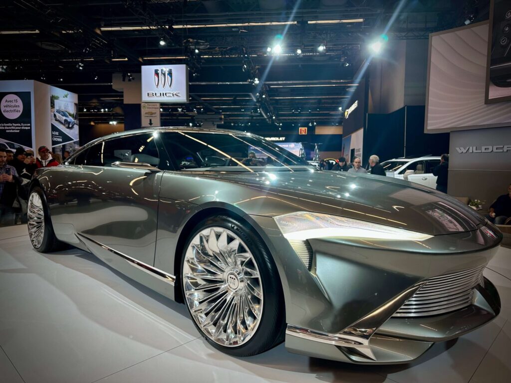 Voiture élégante de couleur vert olive avec des jantes stylées photographiée de près dans une salle de convention. Une enseigne Buick est suspendue en arrière-plan.