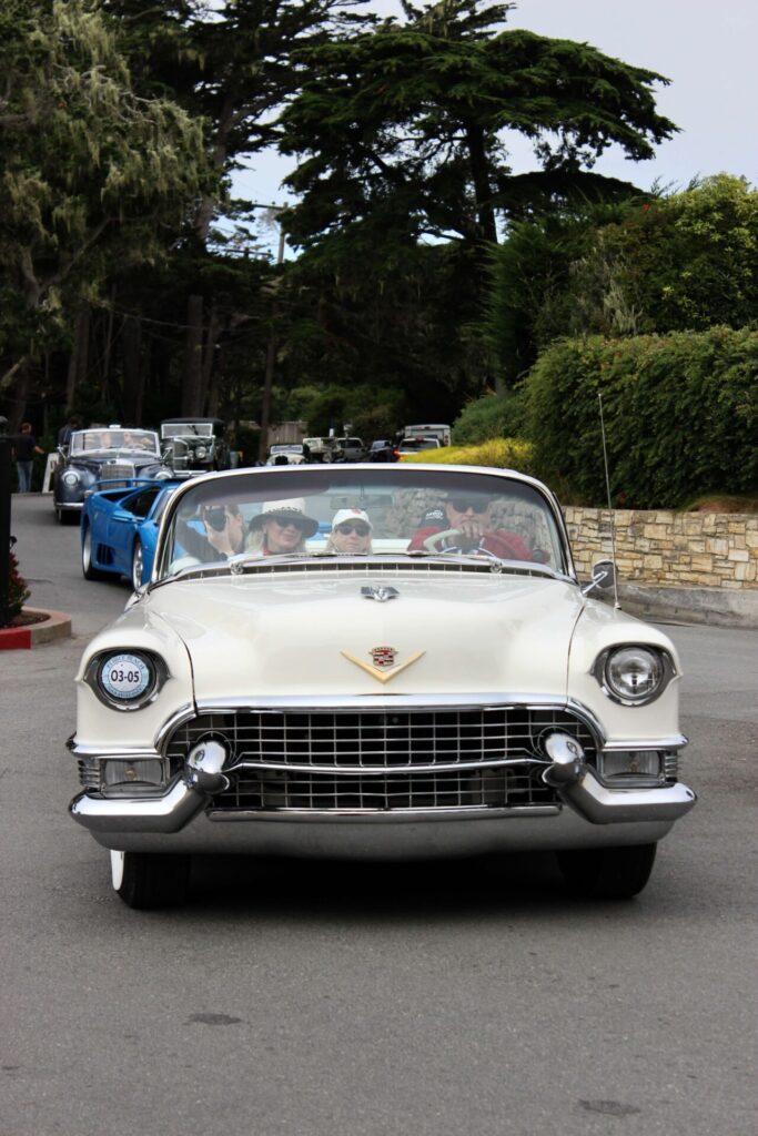 Trois personnes sont photographiées à bord d'une Cadillac blanche qui semble dater des années 1950. La voiture est face à la caméra, et d'autres voitures semblent la suivre en file.