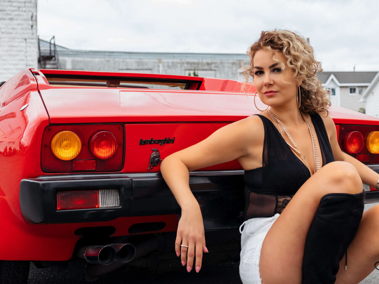 Une fille aux cheveux bouclés est assise, appuyée contre l'arrière d'une voiture de sport rouge. Le nom Jalpa et le logo de Lamborghini sont visibles à côté de son bras droit.