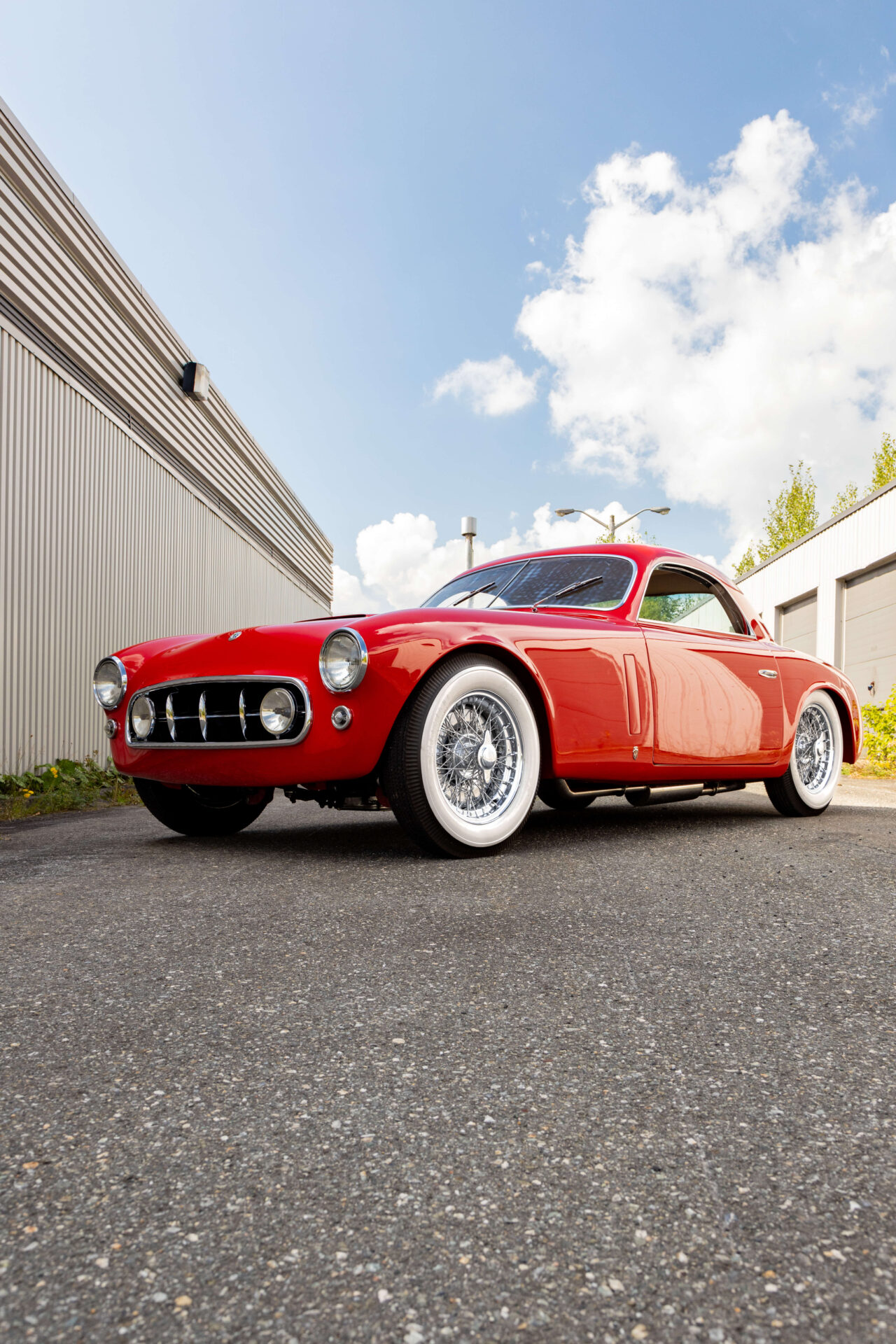 An Alfa Romeo model 6C 2500 in red. The car is pictured on cement and in perspective between a grey building and a row of garage doors.