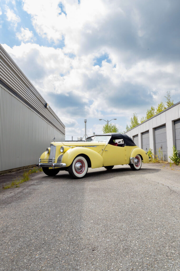 Packard Darrin jaune avec une carrosserie de type Victoria, disposée en angle et en perspective devant des portes de garage à droite et un mur gris uni à gauche.