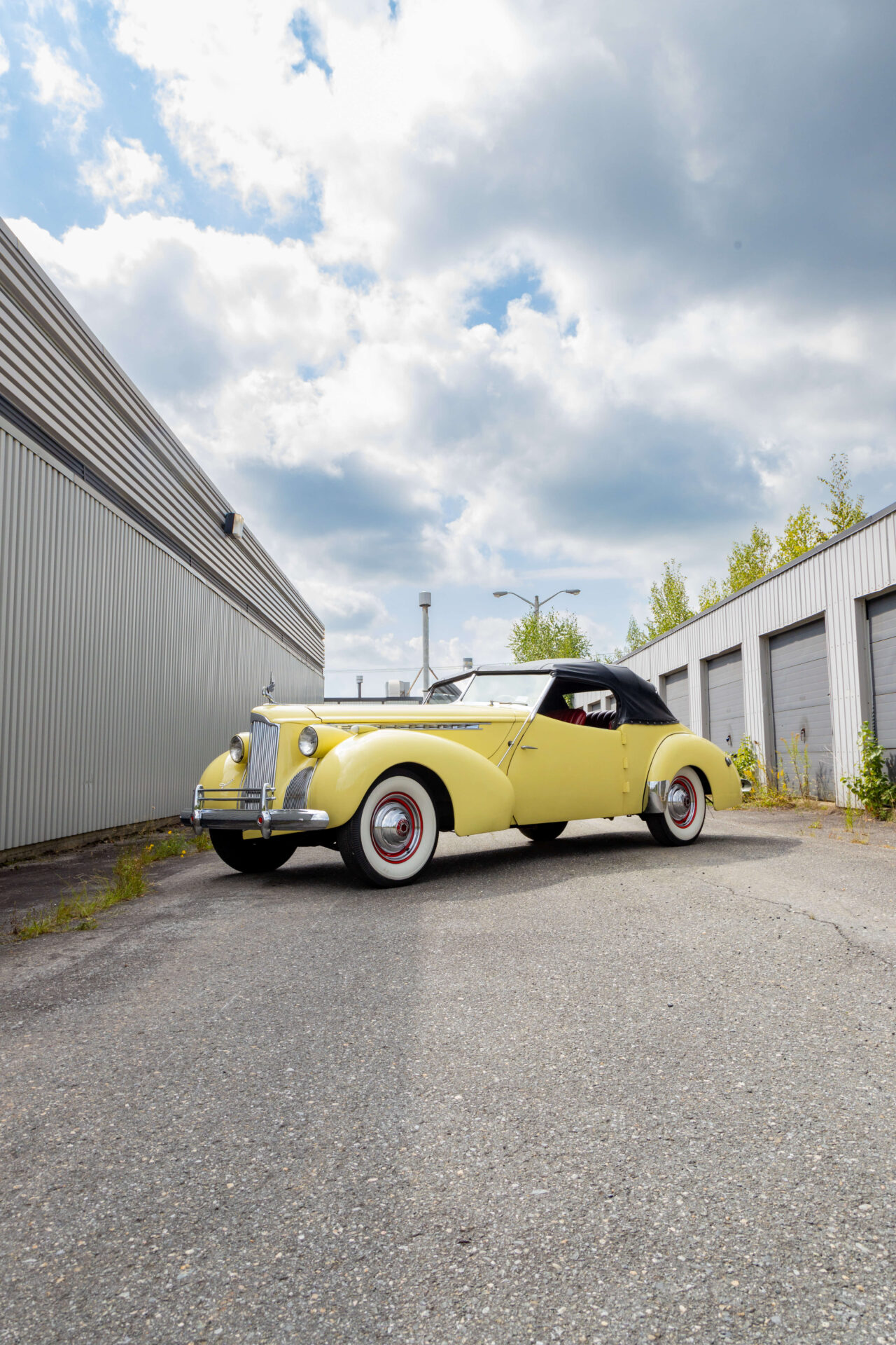 Yellow Packard Darrin with a Victoria body posed at an angle in perspective in front of garage doors on the right and a plain grey wall on the left.