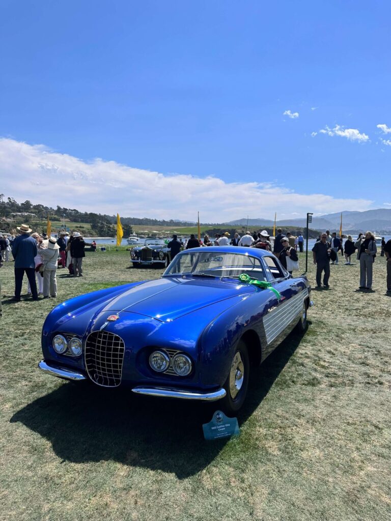 
Cadillac bleue au style distinctif sur l'herbe, entourée de personnes. Une pancarte avec le nom de la voiture est visible près de la roue, et un ruban repose sur le capot de la voiture.