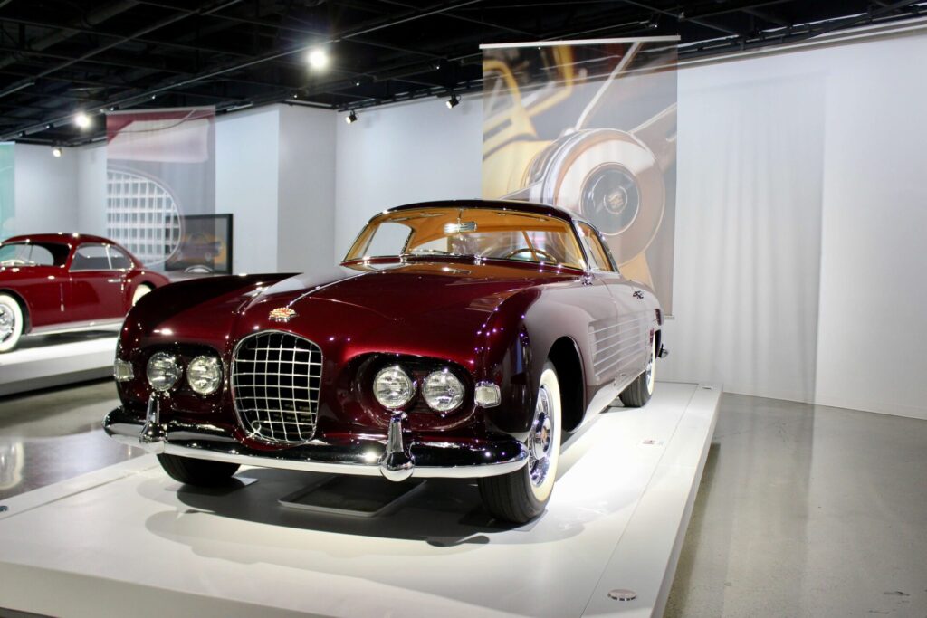 
Un Cadillac Série 62 de couleur aubergine au style distinctif de Ghia. La voiture est sur un socle blanc dans un musée.