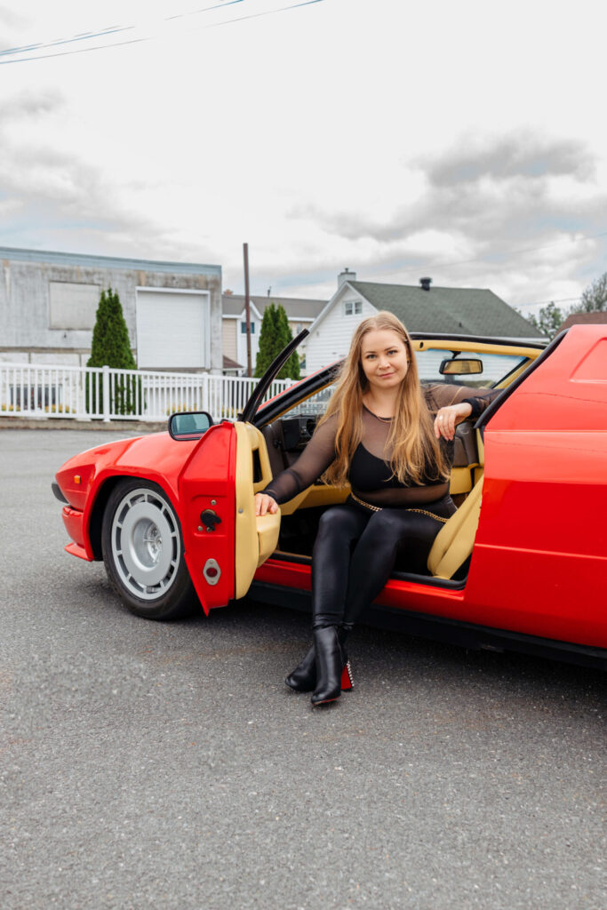 Une fille aux cheveux blonds longs, vêtue de vêtements noirs ajustés, est assise sur le siège avant d'une voiture de sport rouge, les jambes croisées à l'extérieur de la porte ouverte, regardant droit vers l'objectif de la caméra.