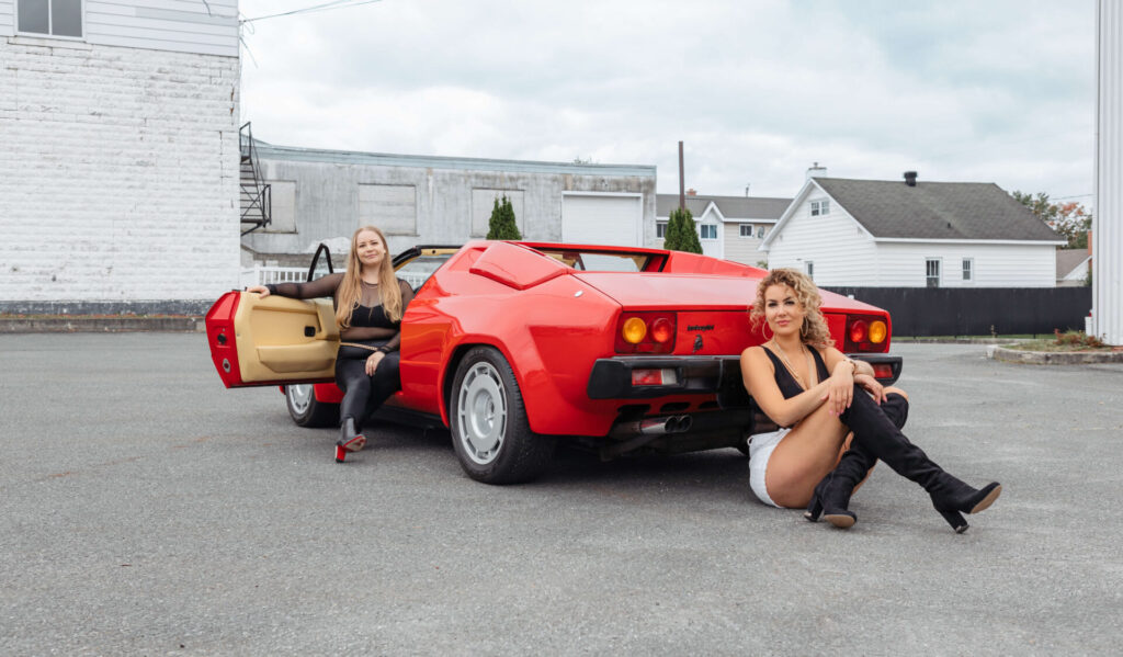 
Deux filles vêtues presque entièrement en noir sont photographiées avec une voiture de sport rouge. L'une est assise au volant avec la porte ouverte, regardant vers l'arrière de la voiture vers la caméra, tandis que l'autre est assise par terre à l'arrière de la voiture, s'appuyant sur la carrosserie.