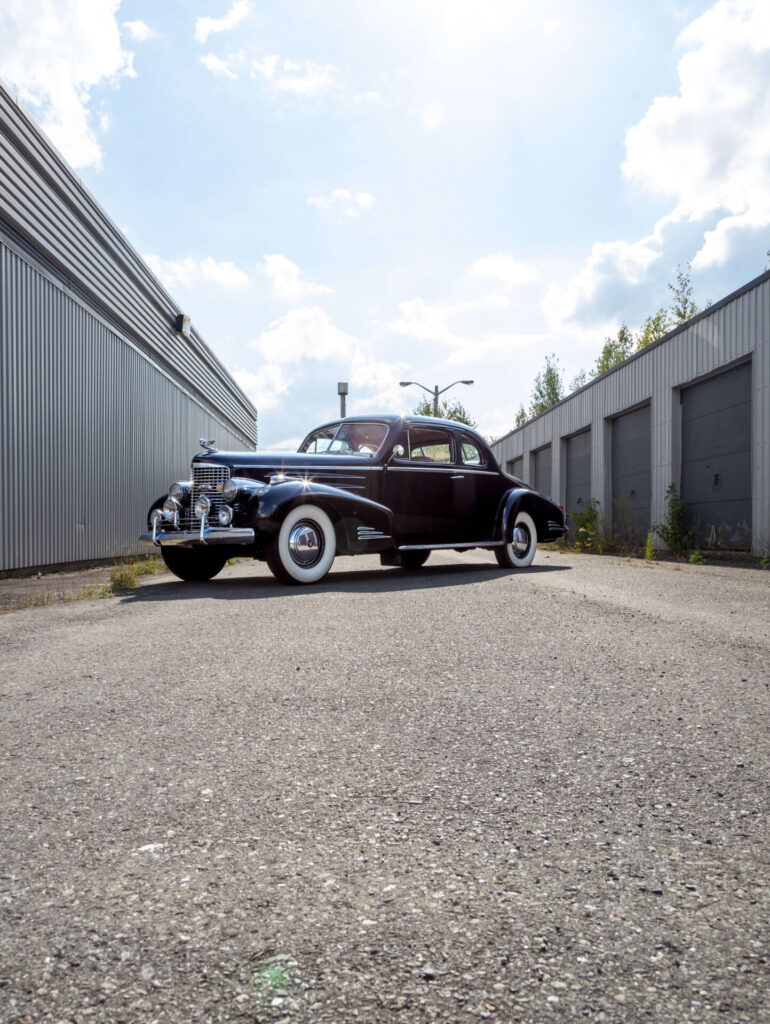 A black 1940 Cadillac V16 is pictured in perspective between a grey building and a row of garage doors.