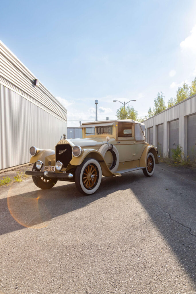 Yellow car with white-flanked tires on wooden wheels. This Pierce-Arrow has a cabriolet roof and a pair of spare tires mounted on the front flanks.