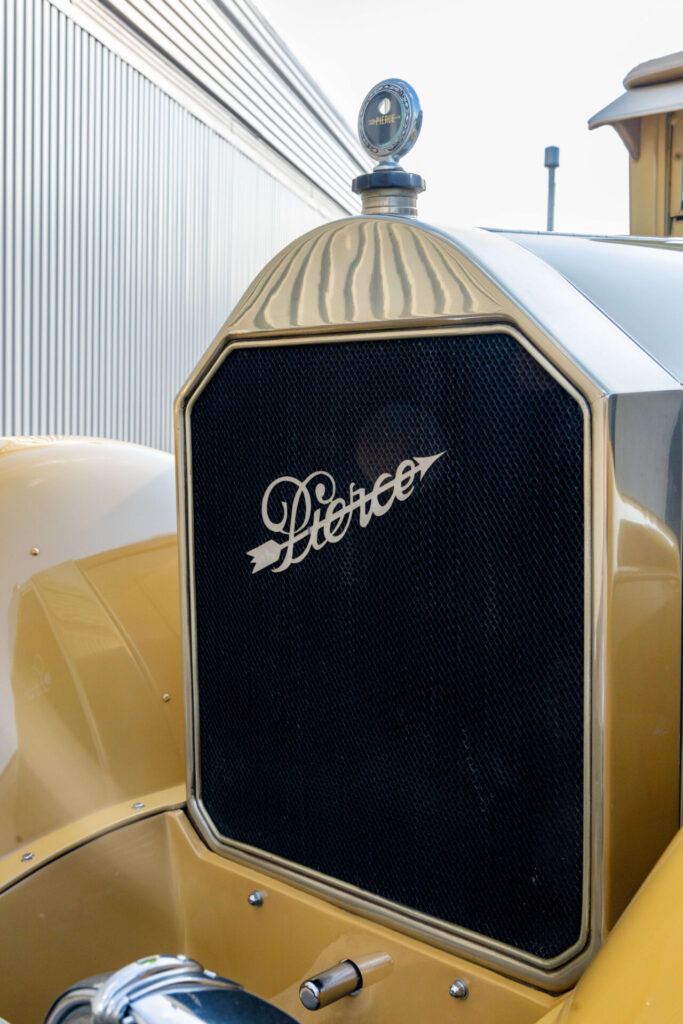 Closer look at the black grille of a yellow, classic car. The word "Pierce" is badged on the grille with an arrow pierced through it.