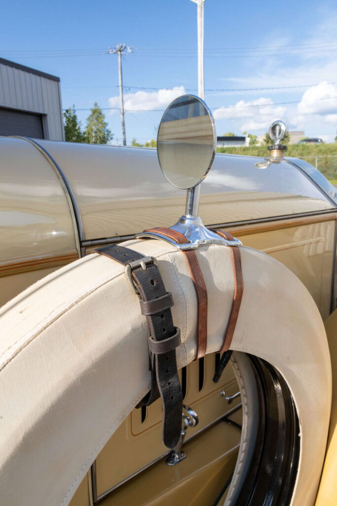 Closer look at a round rear-view mirror mounted by leather straps on the spare tire of a yellow, classic car.