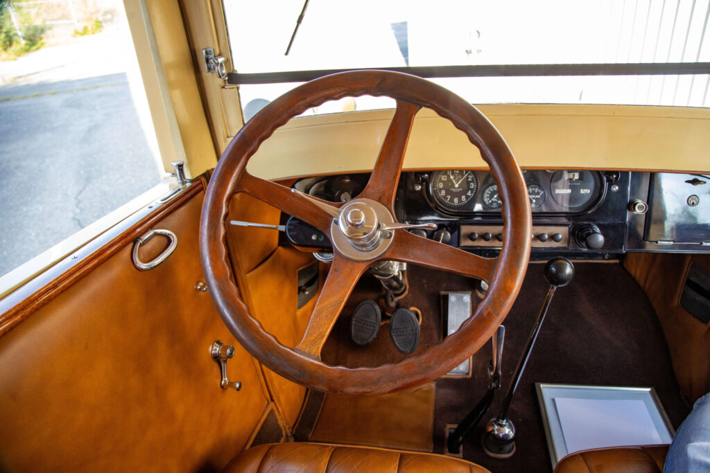 Wooden steering wheel and part of a black dashboard in a classic car. The door panel is visible and made of leather, the same tint as the seat.