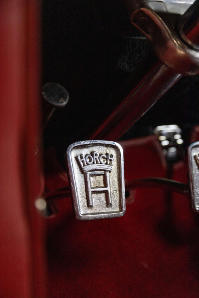 Metal pedal of a classic car. The carpeting is all in red. The Horch logo is moulded on the pedal.