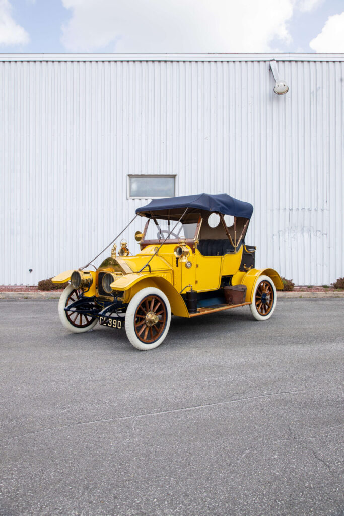 Une Darracq de 1910. Une voiture jaune avec des détails en laiton sur des roues à rayons en bois équipées de pneus entièrement blancs.