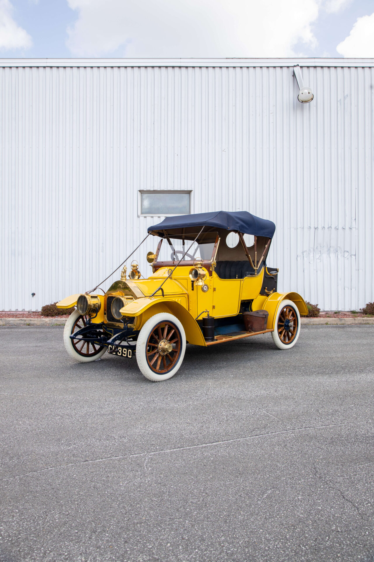 A 1910 Darracq. Yellow car with brass details on wooden-pole wheels with fully white tires.