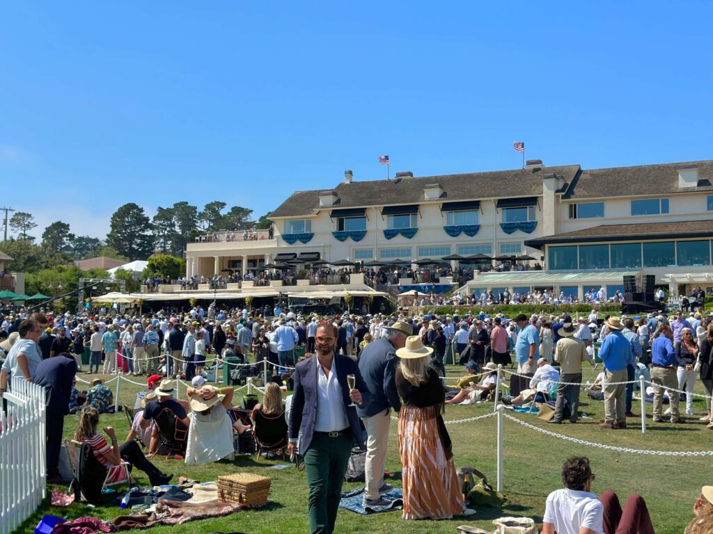 Des gens bien habillés se rassemblent sur l'herbe au Concours d'élégance de Pebble Beach. Certains sont assis sur l’herbe, d’autres debout.