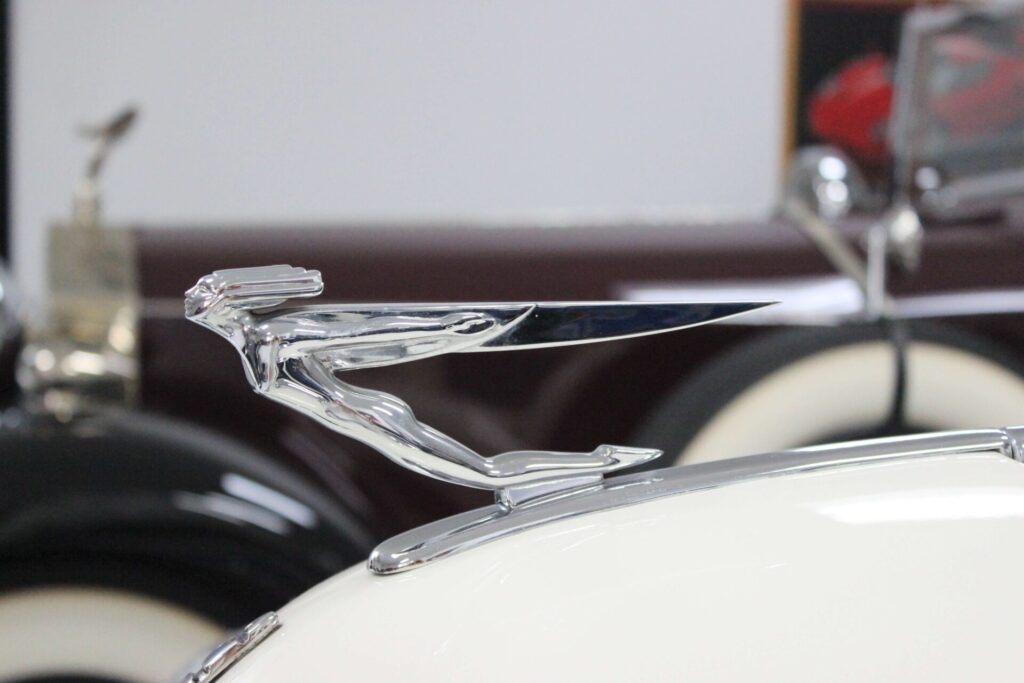 Profile of a hood ornament on a white car. It is a stylised figure of a woman with outstretched arms and hair in the wind.