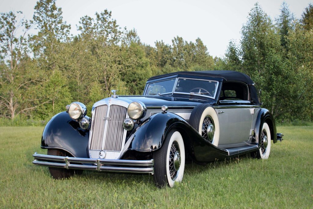 Horch 853. Black and grey classic car viewed at an angle on grass with trees in the background. The car has a silver-coloured angled grille, white flanked tires on all wheel and on the spare wheel mounted on the flank of the front wing, the windshield is divided in two sections and the convertible top is black.