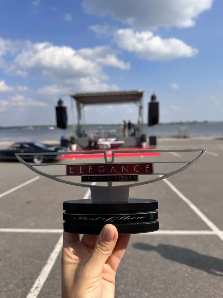 Une troupe de jury sur une scène dans un parking avec le fleuve et le ciel en arrière-plan. Le trophée ressemble à deux ailes stylisées et on peut lire dessus les mots « Élégance Trois-Rivières