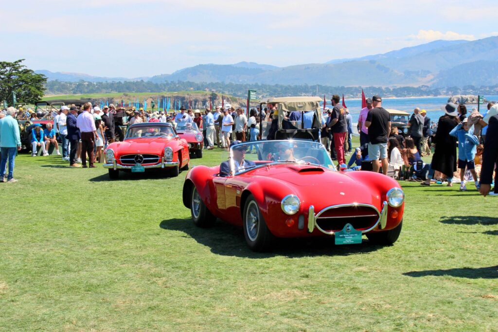 Deux voitures rouges roulent l'une derrière l'autre sur l'herbe devant un terrain au Concours d'Élégance de Pebble Beach