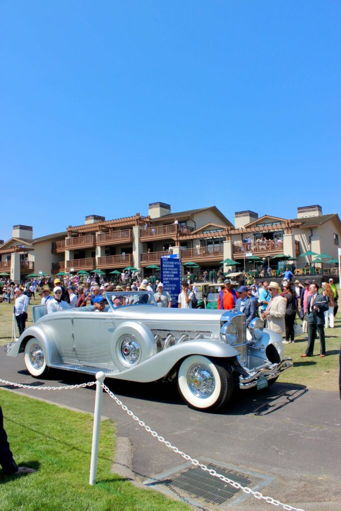 Une voiture argentée roule sur une route devant un imposant hôtel. Il y a du monde partout y compris sur tous les balcons de l'hôtel. C'est la cérémonie au Concours d'Élégance de Pebble Beach.