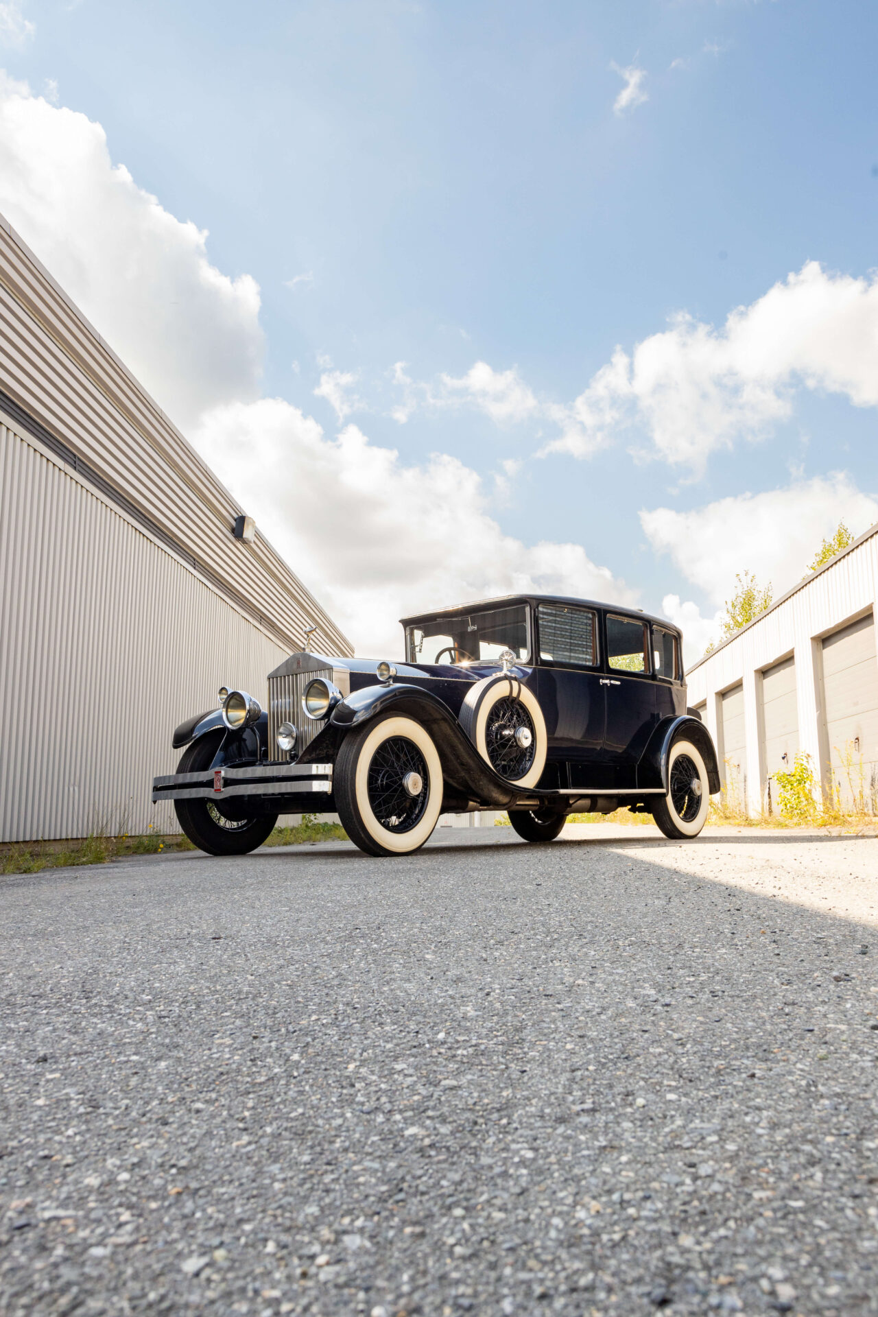 A dark blue car pictured in perspective on asphalt between two grey building and a blue sky with little clouds. The car is imposing and has white-flanked tires. A spare tire is visible on the running board.