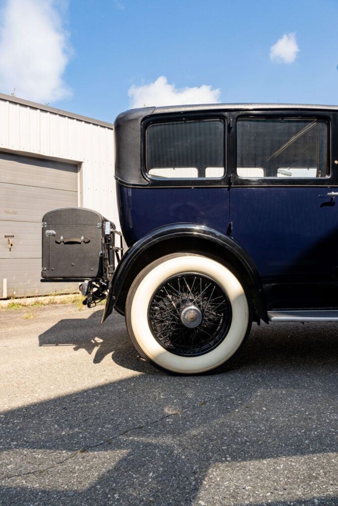 Rear end of a dark blue car seen from the side. The rear wheel has a white-flanked tire and a separate black luggage trunk is attached to the back of the car.