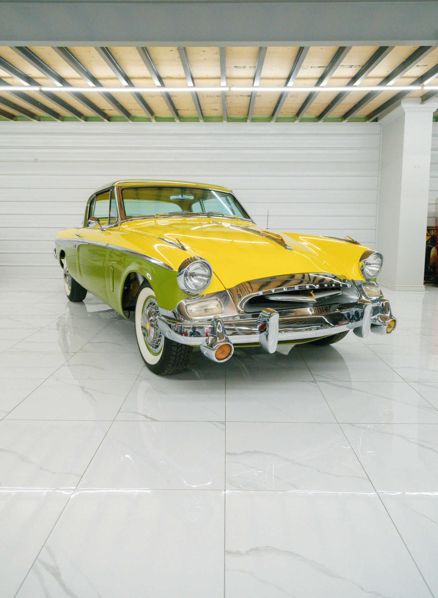 Yellow and lime-green car from the 1950s. The Studebaker President Speedster is pictured here at an angle from the front on a white marble floor.