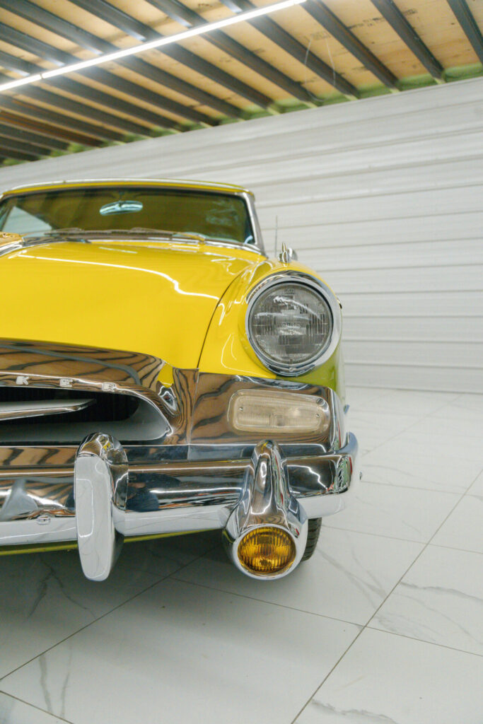 Vue partielle avant d'une voiture jaune et verte. La voiture des années 1950 possède un très gros pare-chocs chromé.