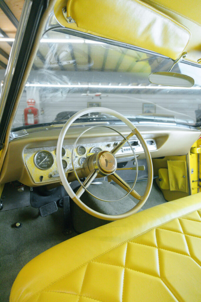 View of the dashboard and steering wheel of a car from the 1950s. The leather seat is yellow.