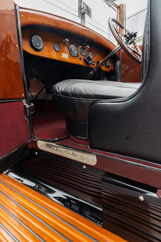 Closer look through the open passenger door of an old car. The dashboard is made of wood and the seat is in black leather. A metal badge states the car's coachbuilder: Halls, Lewis & Co