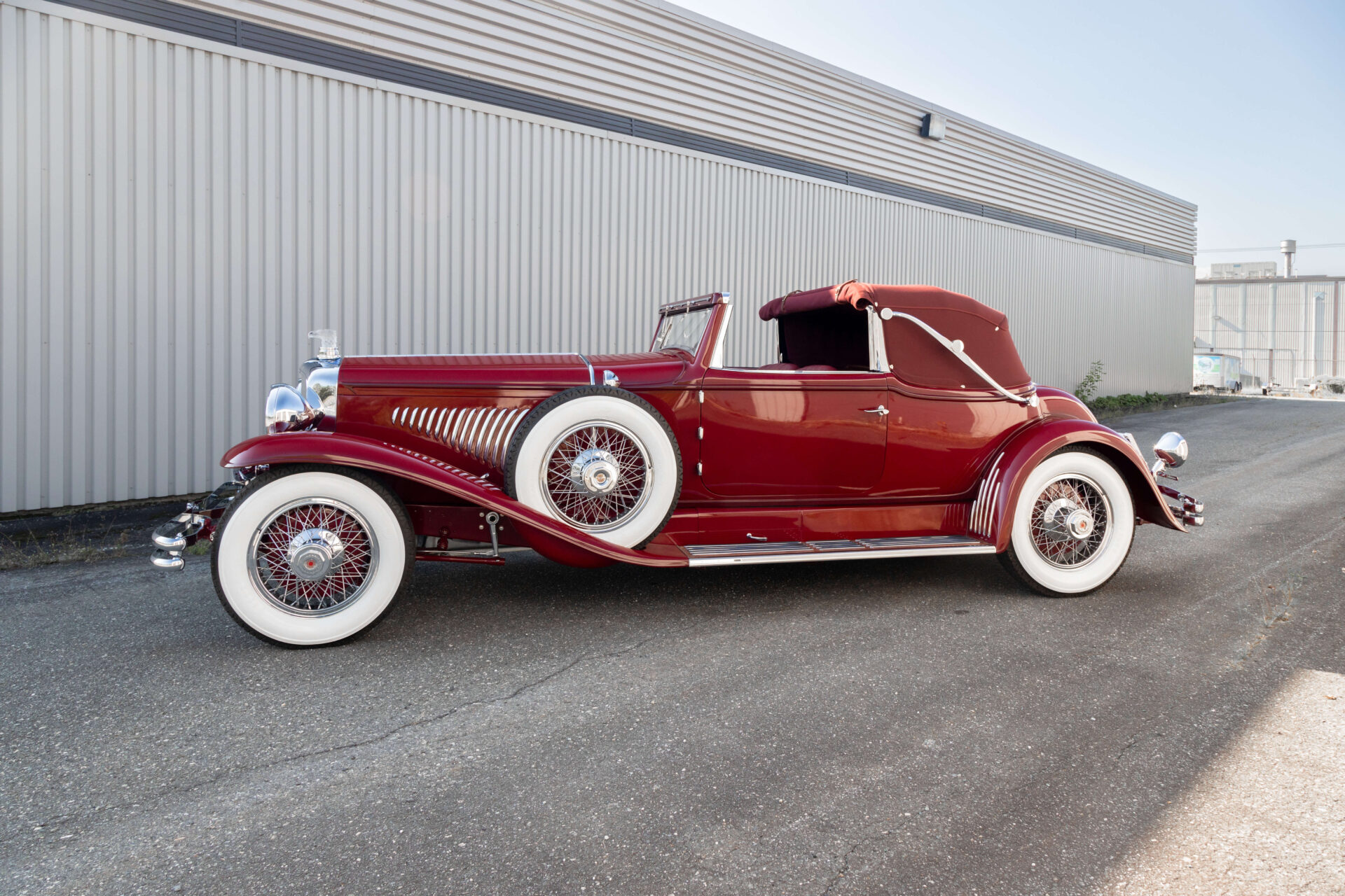 Profil view of a red classic car, a Duesenberg Model J. It is a cabriolet style and a spare wheel is mounted on its front fender. All three wheels visible have white-flanked tires