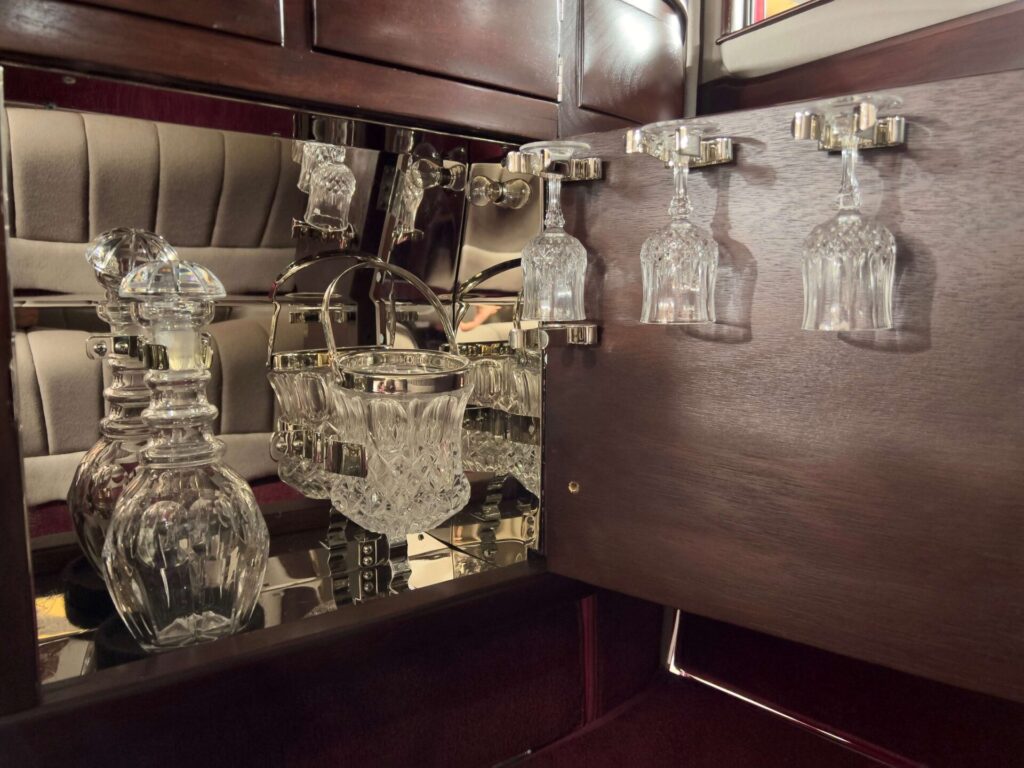 A wooden bar cabinet with one door open showing three Chrystal glassed hanging upside-down on the door and a carafe and ice bucket in the bar.