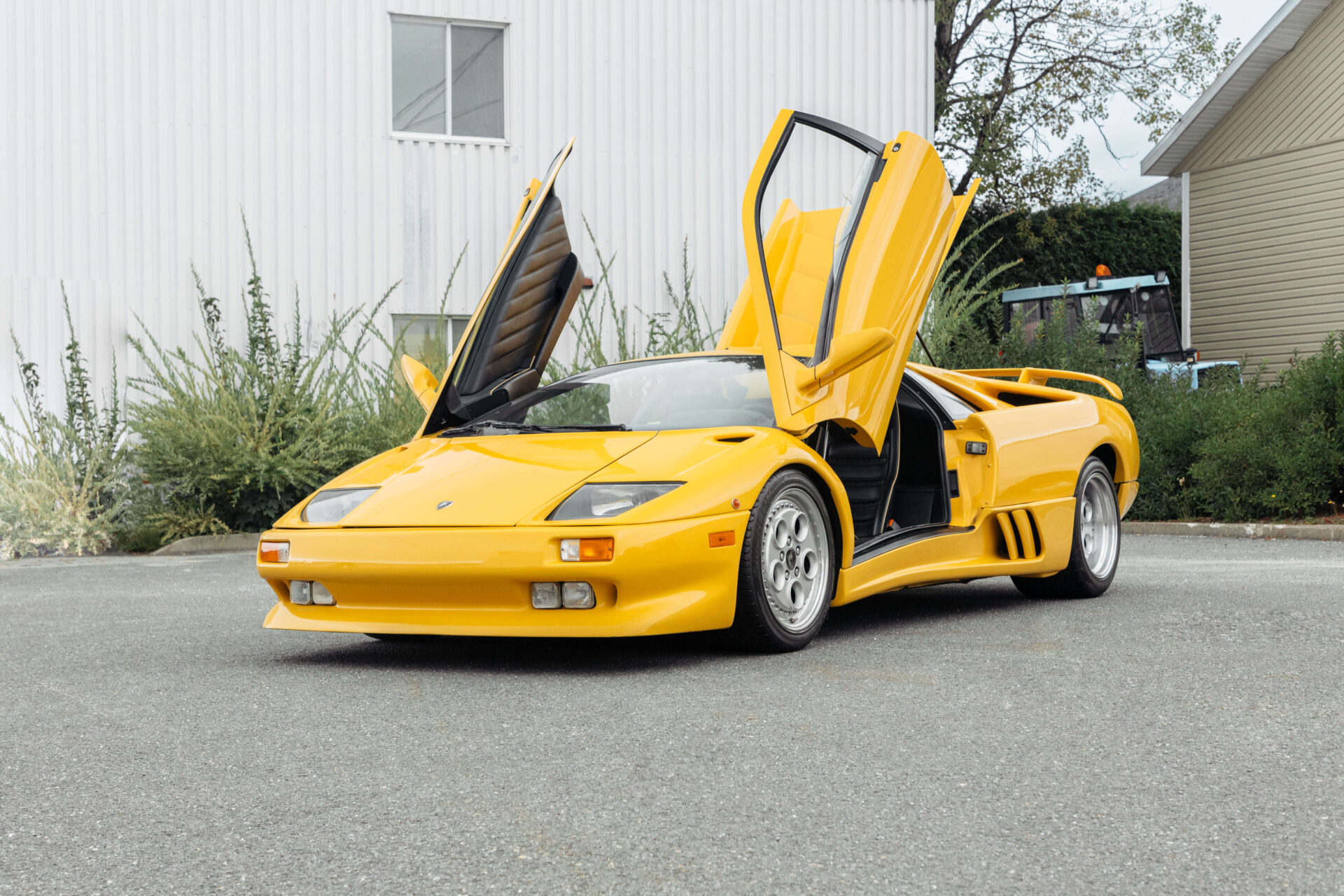 Yellow car with scissors doors open pictured on cement in front of a white building and bushes.