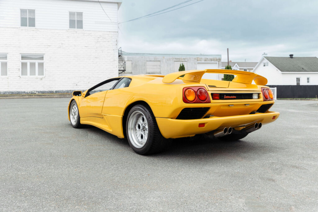 Une supercar jaune photographiée en angle depuis l'arrière. La voiture est large et basse. Un bâtiment industriel peut être vu en arrière-plan.