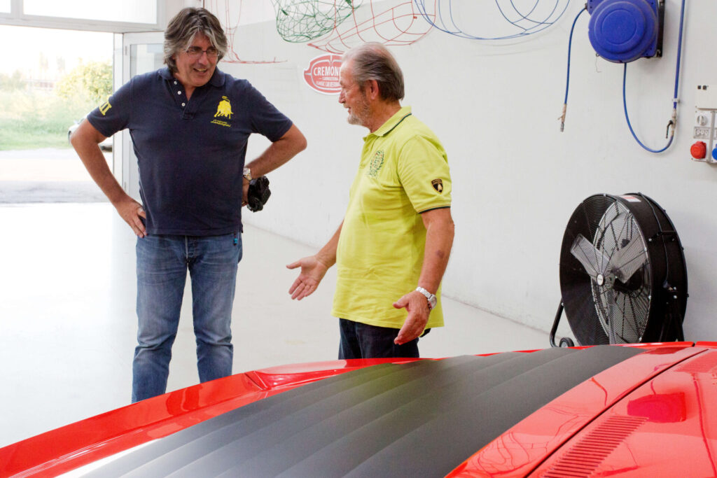 Deux hommes sont debout à discuter devant un mur blanc de garage et derrière une voiture rouge.